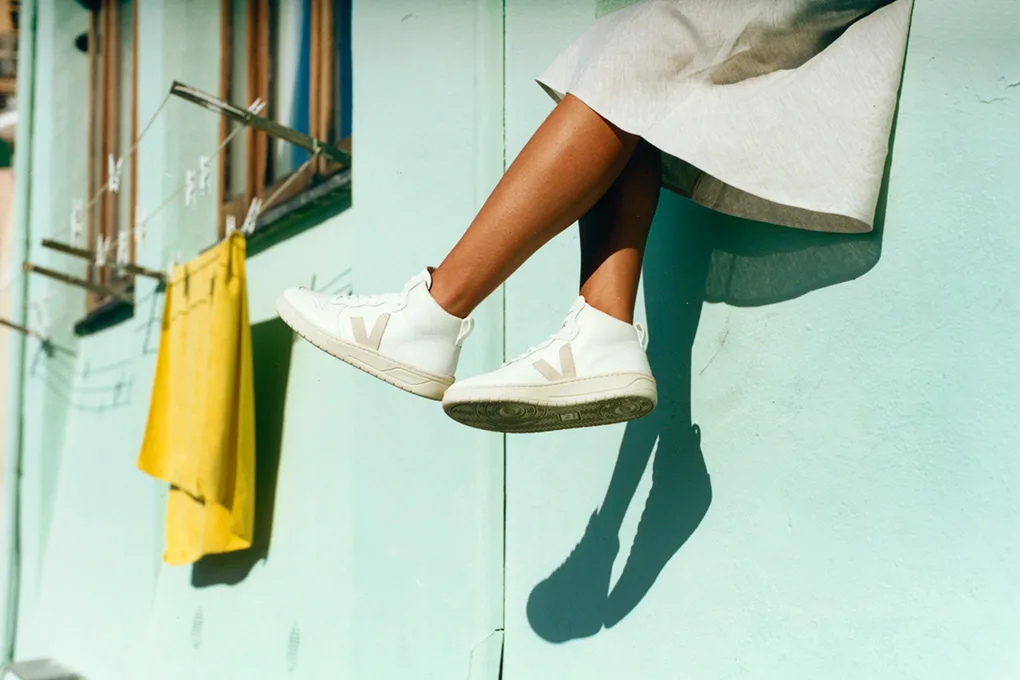 Eine Frau lässt ihre Füße mit Veja-Sneakern vor einer türkisen Hauswand baumeln